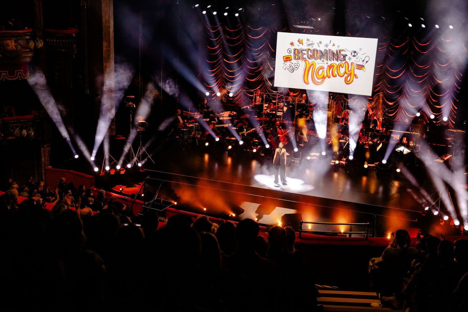 Concert stage with dramatic white spotlights and large projection screen visible above the audience.