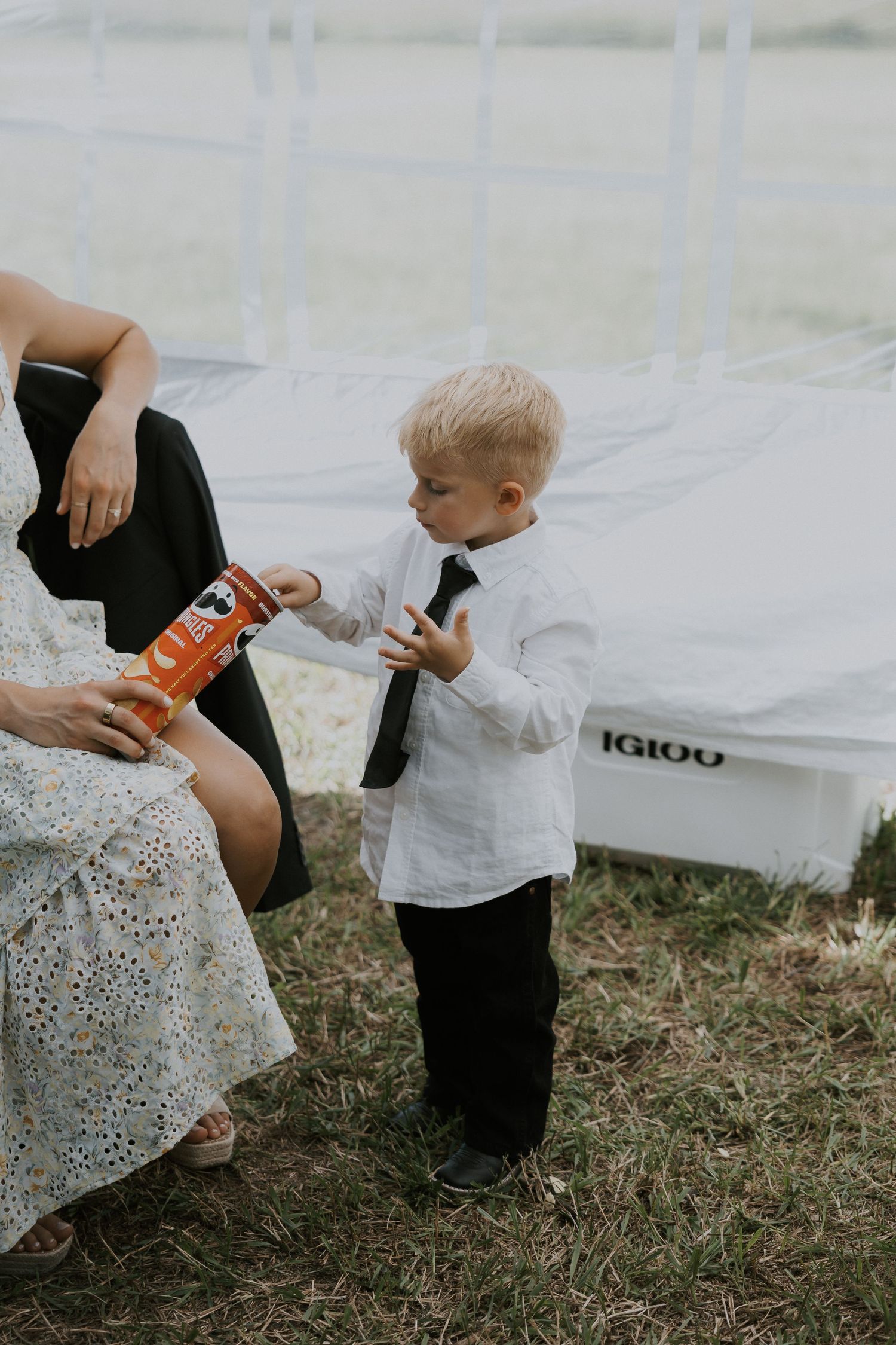 Small child in white shirt stands outdoors holding a drink near an Igloo cooler while an adult holds another beverage.