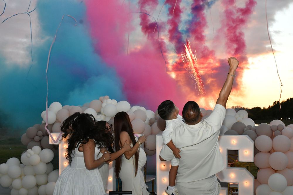 Group of people celebrating at sunset with pink smoke bombs and white balloon decorations creating a festive atmosphere.