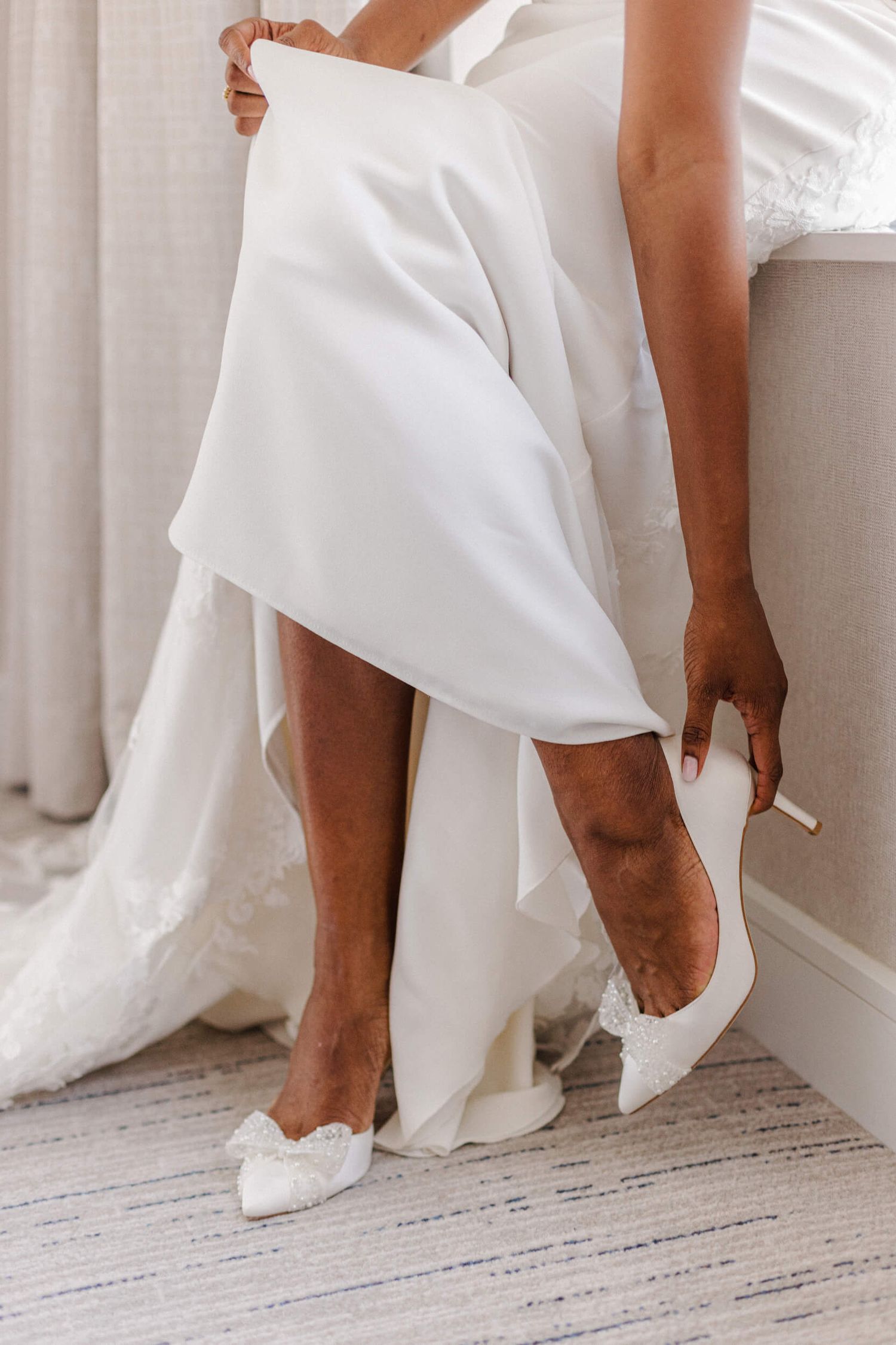 Close-up view of elegant white bridal shoes and flowing wedding dress hem on light wooden floor.