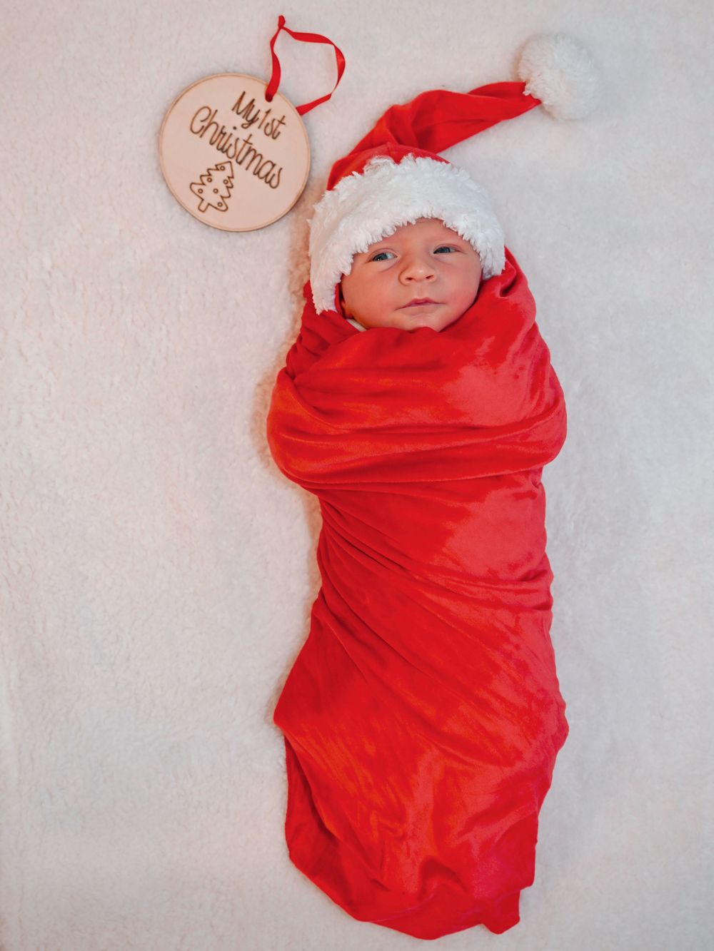 Newborn baby swaddled in red blanket wearing a Santa hat beside a My First Christmas ornament.