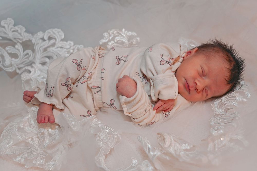 Sleeping newborn in a delicate floral onesie resting on white lace fabric.