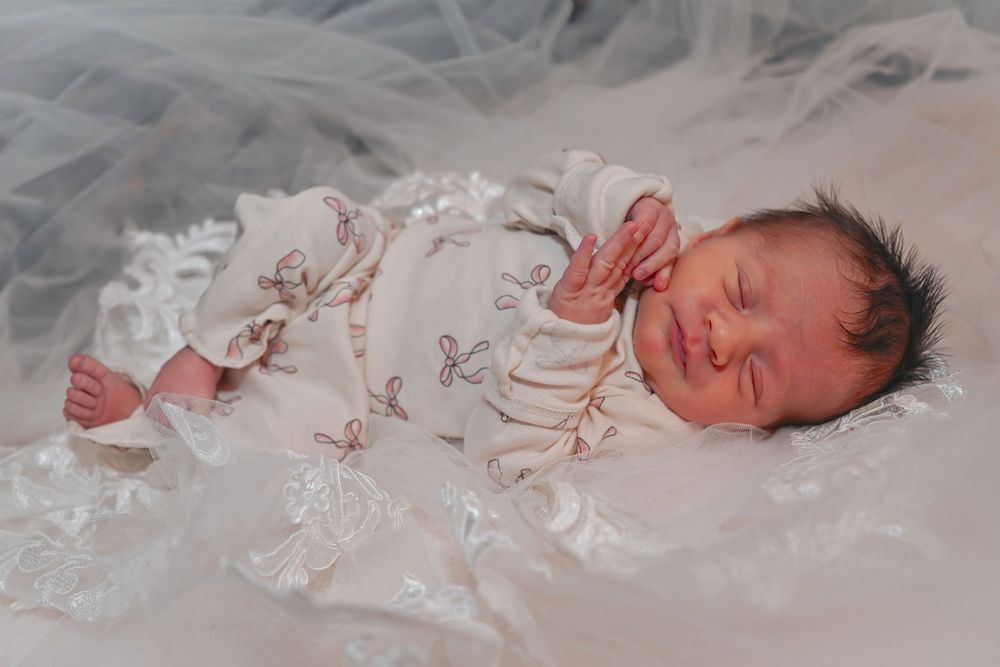 Peaceful newborn baby sleeping in a white floral outfit on soft white lace fabric.