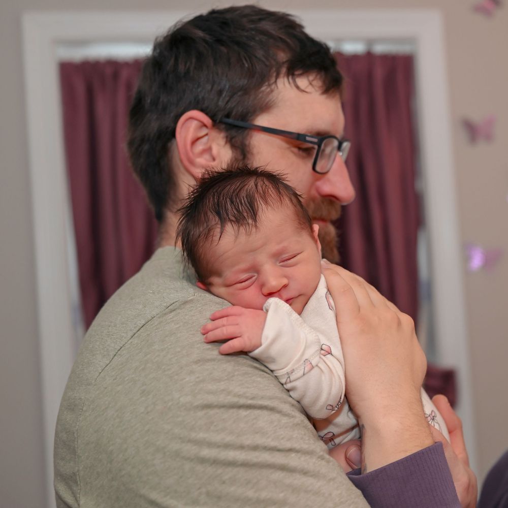 Father with glasses tenderly holding his sleeping newborn baby against his shoulder.