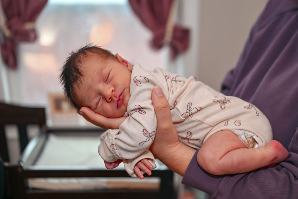 Newborn baby sleeping peacefully in an adult's hands wearing a white patterned outfit.