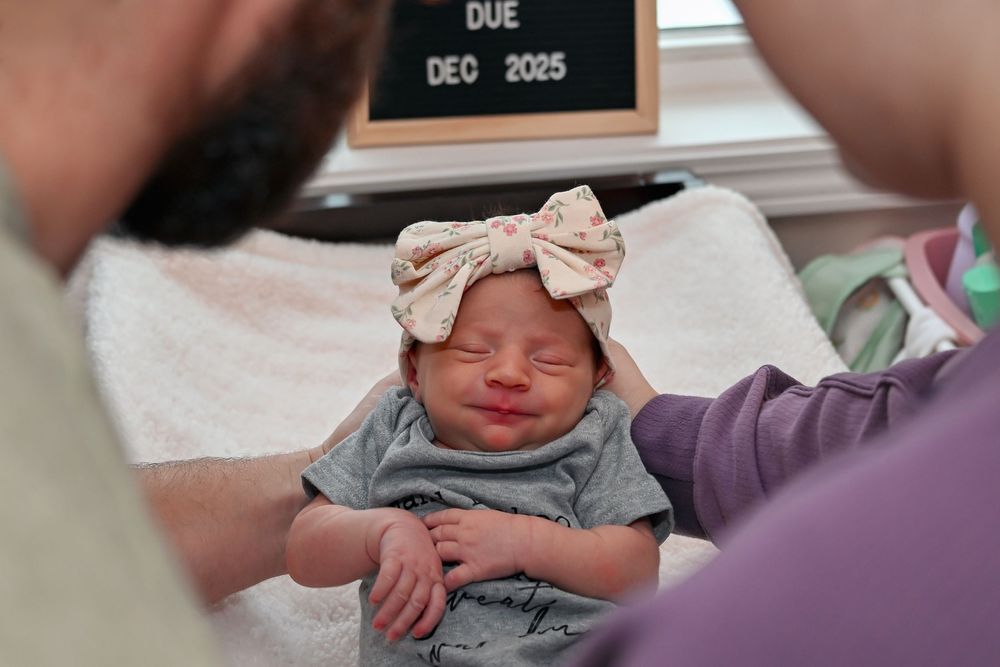 Newborn baby girl with a bow headband smiling with a due date sign in the background.
