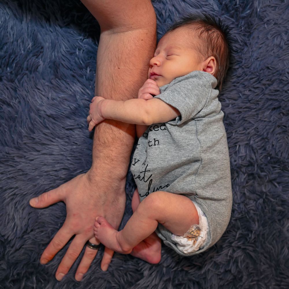 Newborn baby in a gray onesie sleeping beside a parent's arm on a blue fluffy blanket.