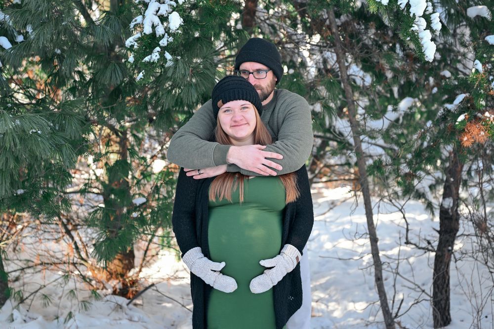 Happy expecting couple embracing outdoors in winter snow, showing baby bump in a green maternity dress.