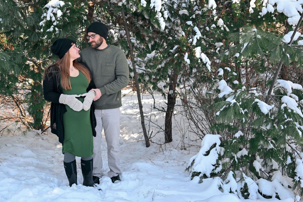 Couple smiling at each other in snowy winter forest, woman in green dress and man in gray sweater, surrounded by snow-covered trees.