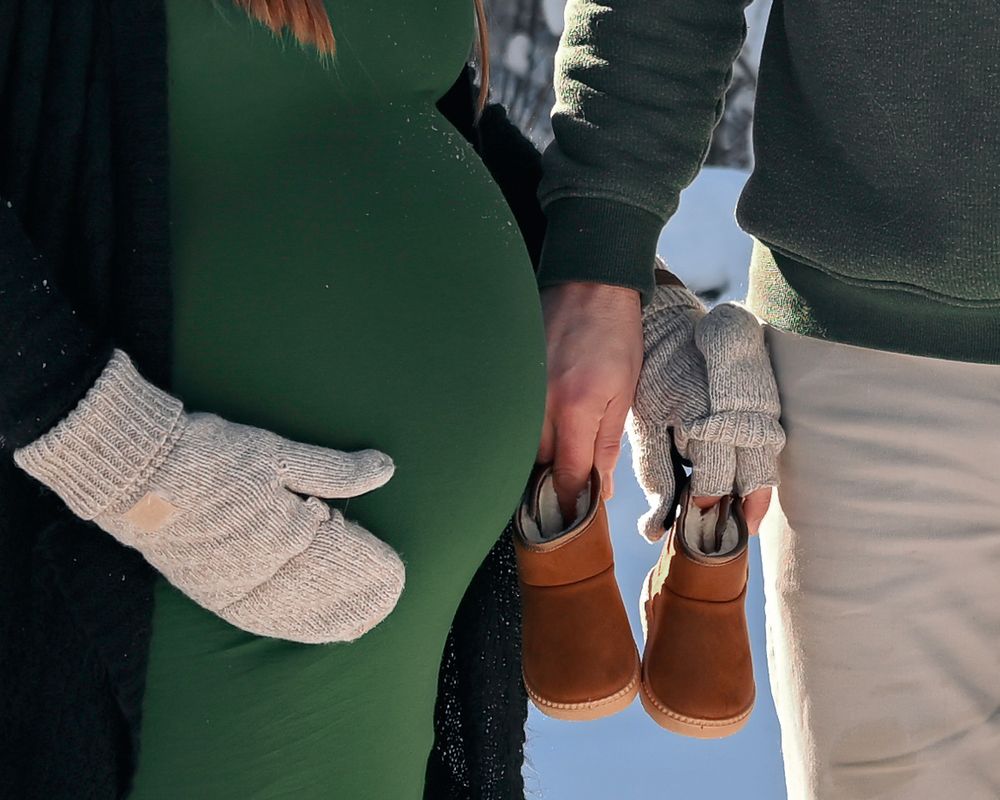 Pregnant couple in winter holding tiny brown baby boots and wearing knit mittens outdoors in snow.