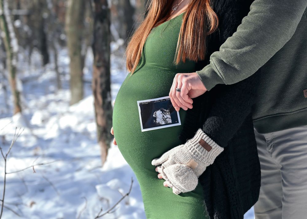 Pregnant couple holding ultrasound photo outdoors in snowy winter forest, woman in green dress wearing knit mittens.
