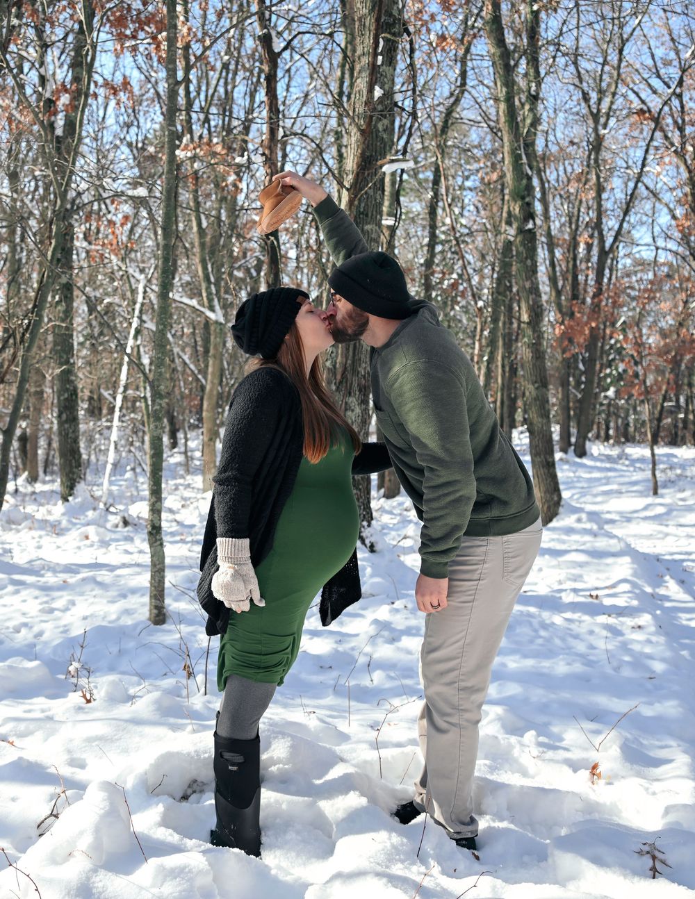 Couple kissing under mistletoe in snowy winter forest, woman in green dress and black boots, man in gray outfit.