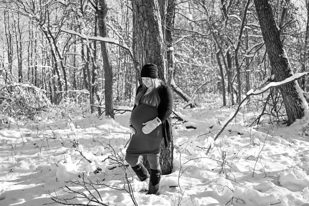 Person walking through snowy winter forest in black and white, wearing dark coat and hat among bare trees.