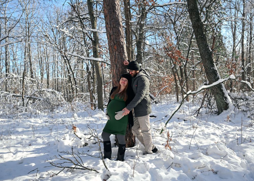 Couple embracing in snowy winter forest, woman in green dress showing baby bump, surrounded by snow-covered trees.