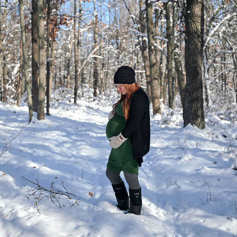 Pregnant woman in green dress and black coat standing in snowy winter forest trail wearing boots and beanie hat.