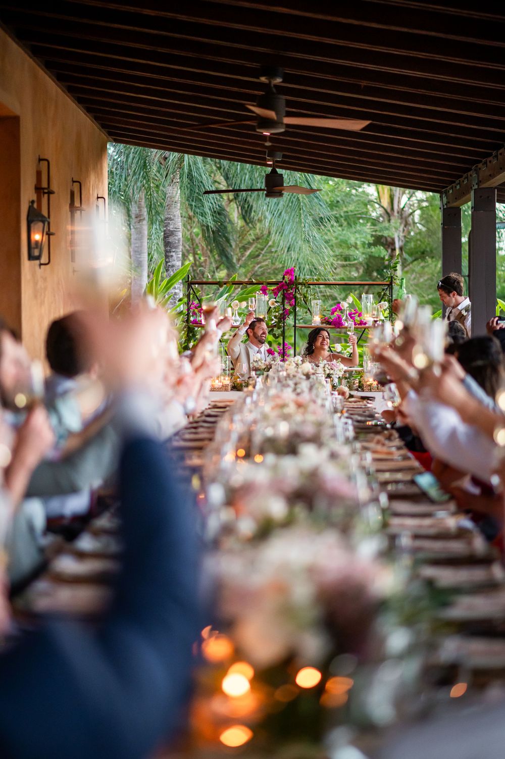 Wedding celebration with guests raising hands in joy at a long outdoor dining table.
