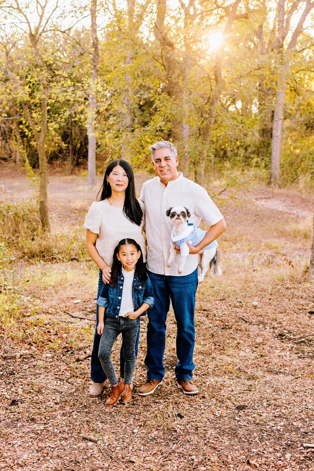 A trio stands together outdoors in a wooded area during golden hour with their small white dog.