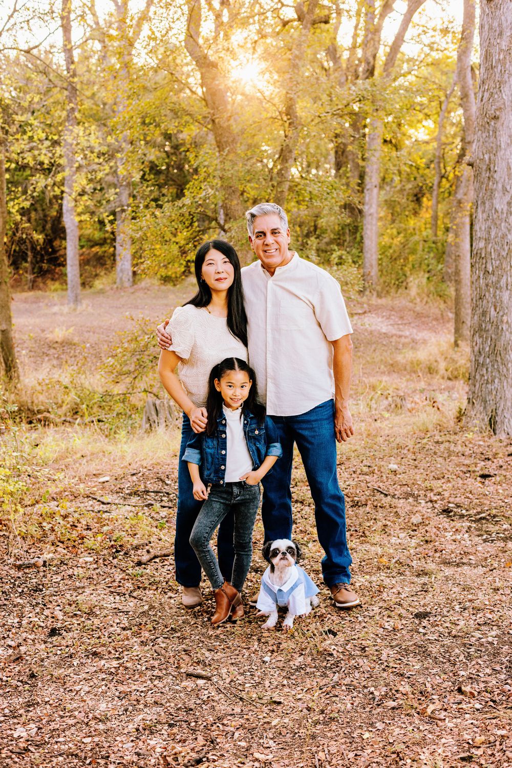 A group stands together with their small white dog in a sunlit wooded area during golden hour.