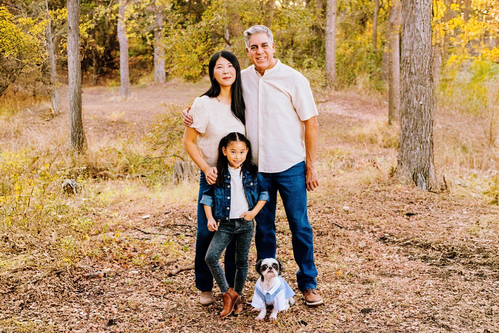 Three people and a small white dog stand together on a forest path surrounded by autumn foliage.