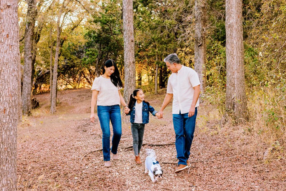 Family walking together with small white dog on autumn trail through wooded area.