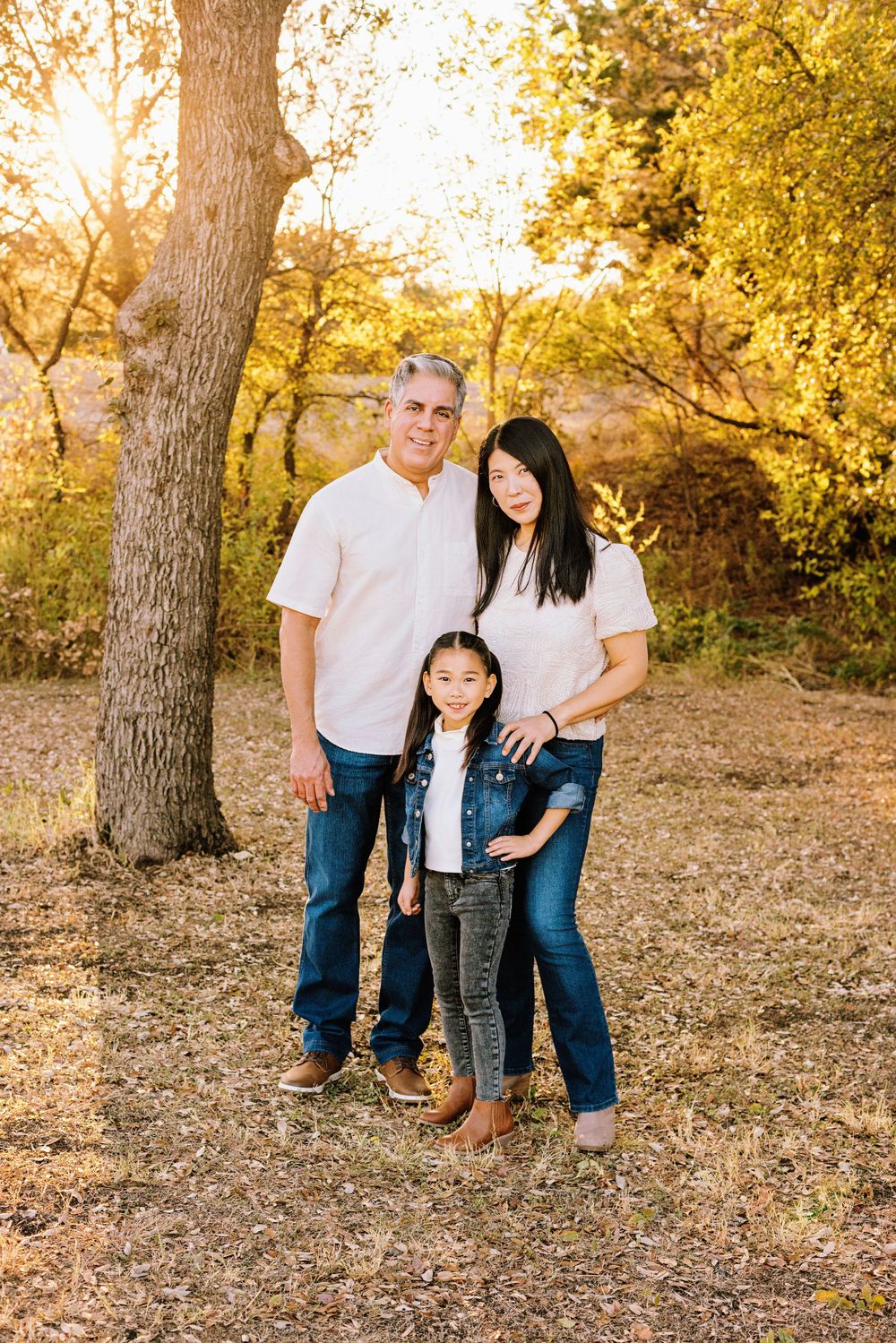 A family in coordinated denim and white outfits poses together in a sun-lit autumn forest setting.
