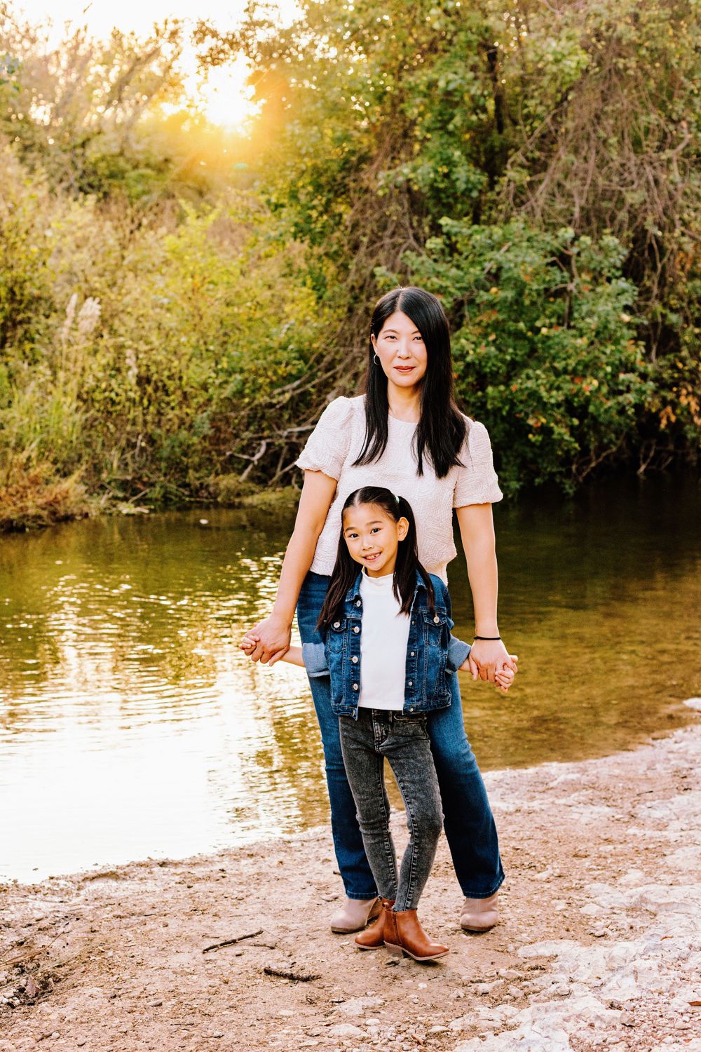 Two people in casual clothing standing by a lake at sunset with golden light filtering through trees in the background.