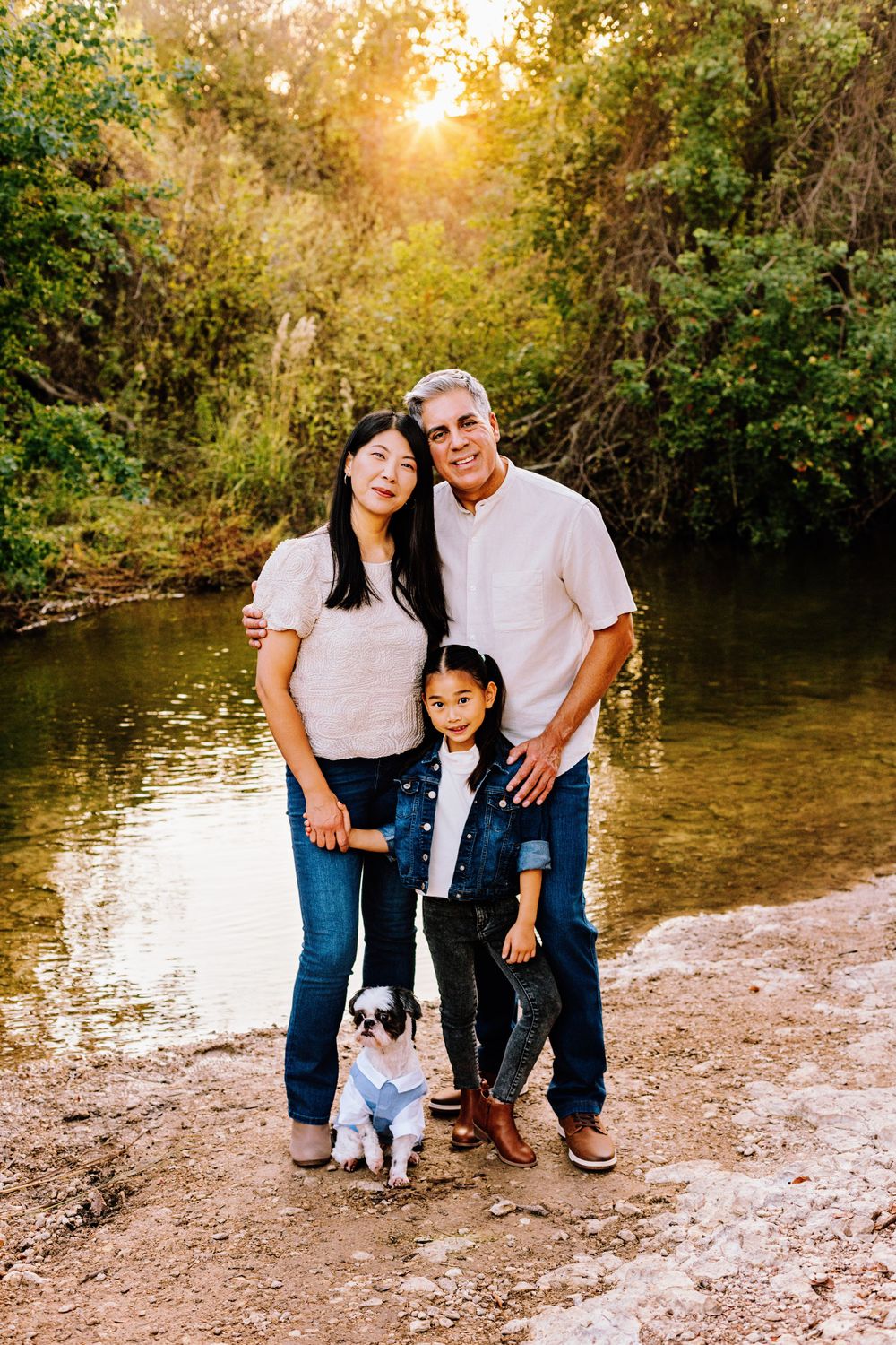 A family poses together at sunset by a creek with their small white dog, surrounded by lush greenery.