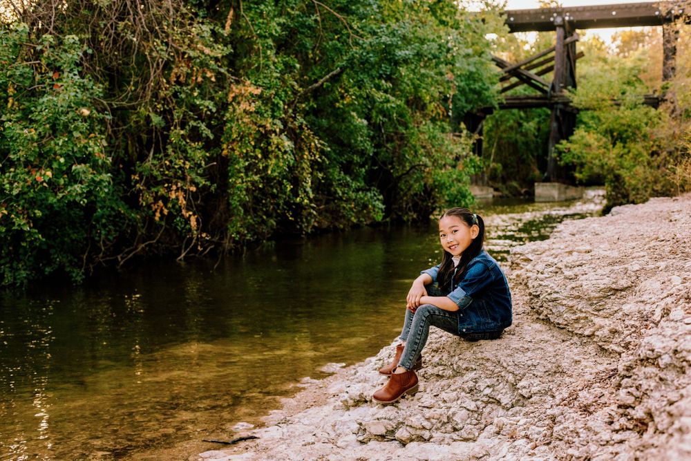 A person in a denim jacket sits on the sandy riverbank beneath an old wooden railway bridge during autumn.