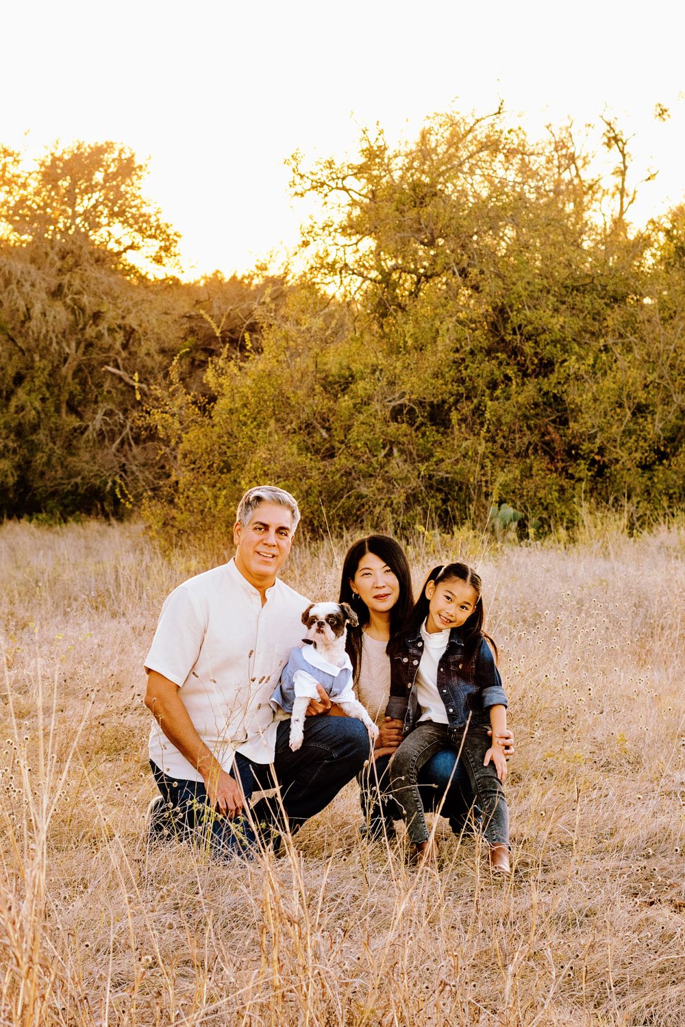 Family with small dog poses together in tall dry grass with sun-dappled trees in the background.