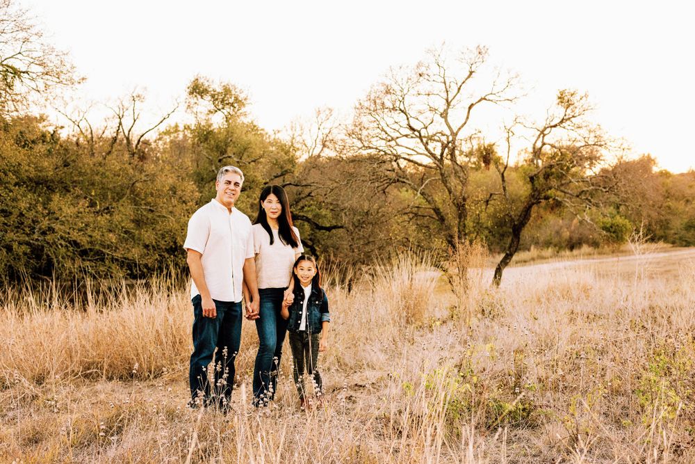 Family poses together in a golden grass field at sunset with trees in the background.