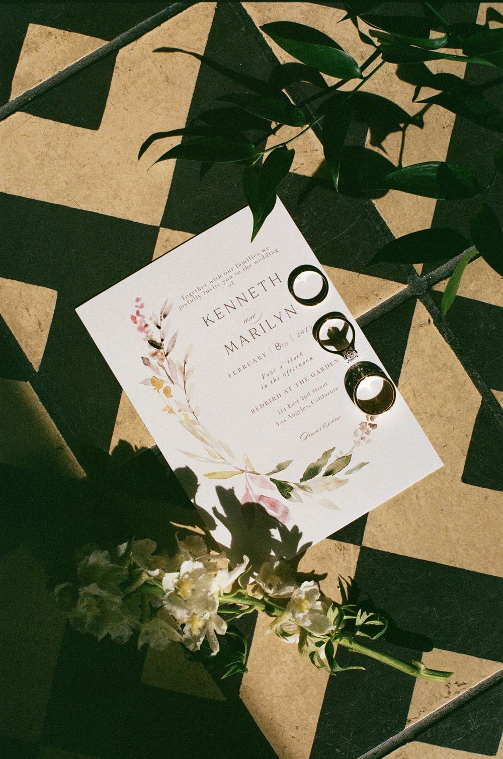 Wedding invitation for Kenneth and Marilyn with floral wreath design, gold rings, and white flowers on geometric tile.