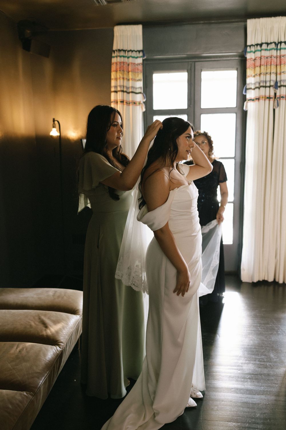 Bridesmaids helping bride put on veil and wedding dress in warmly lit hotel room.