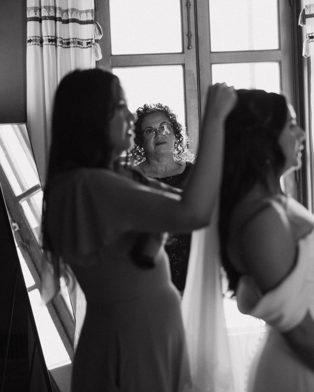 Black and white photo of bridesmaids helping bride with veil near bright window.