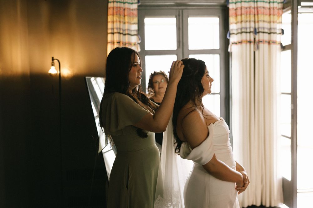 Bridesmaid in green dress helping bride with hair accessories near window in warmly lit room.