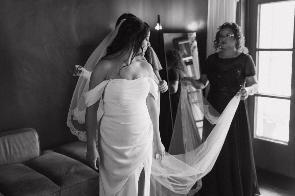 Black and white photo of bride in white gown having veil arranged by helper near window.