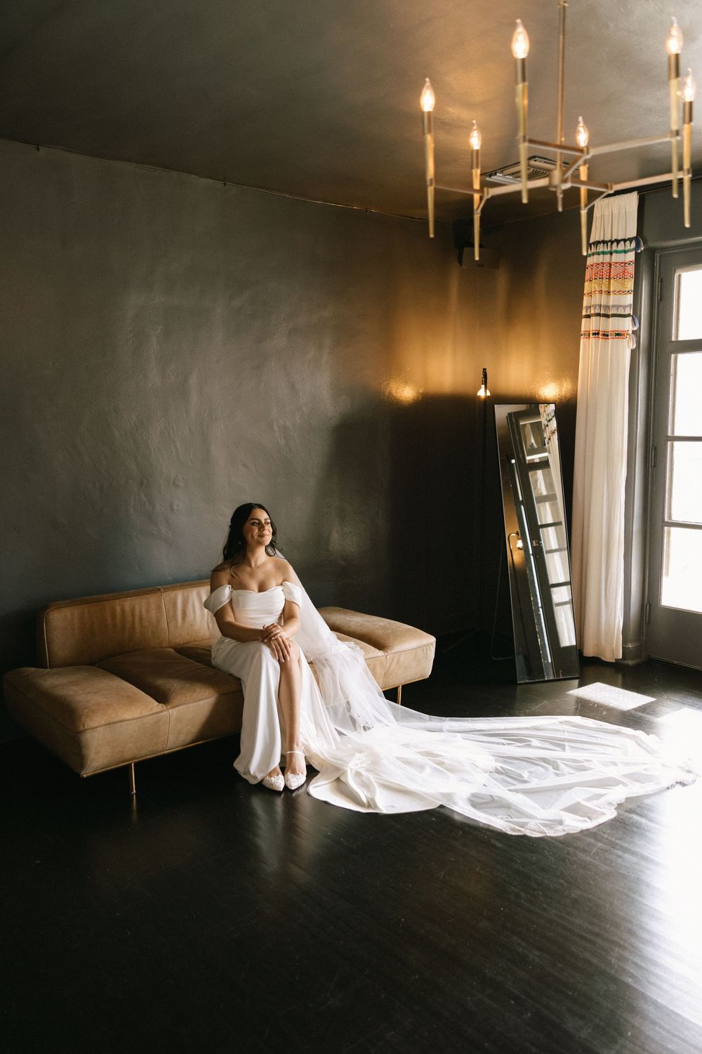 Elegant bride in off-shoulder white gown with long train seated on leather sofa under chandelier.