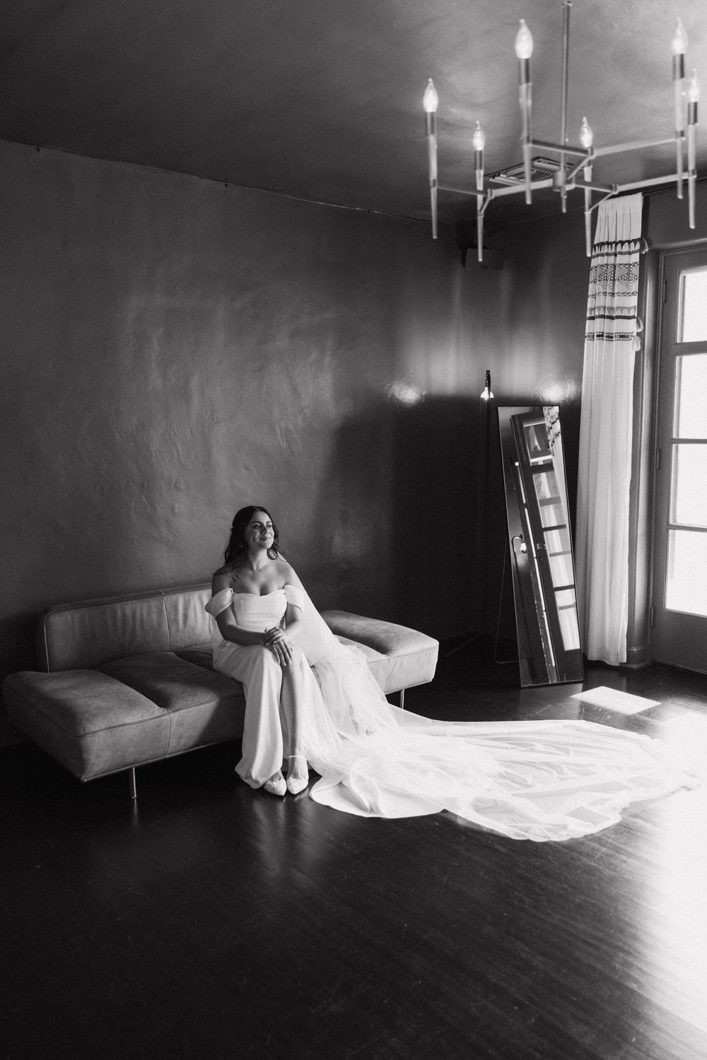Black and white portrait of bride in off-shoulder wedding dress with cathedral veil on leather sofa.