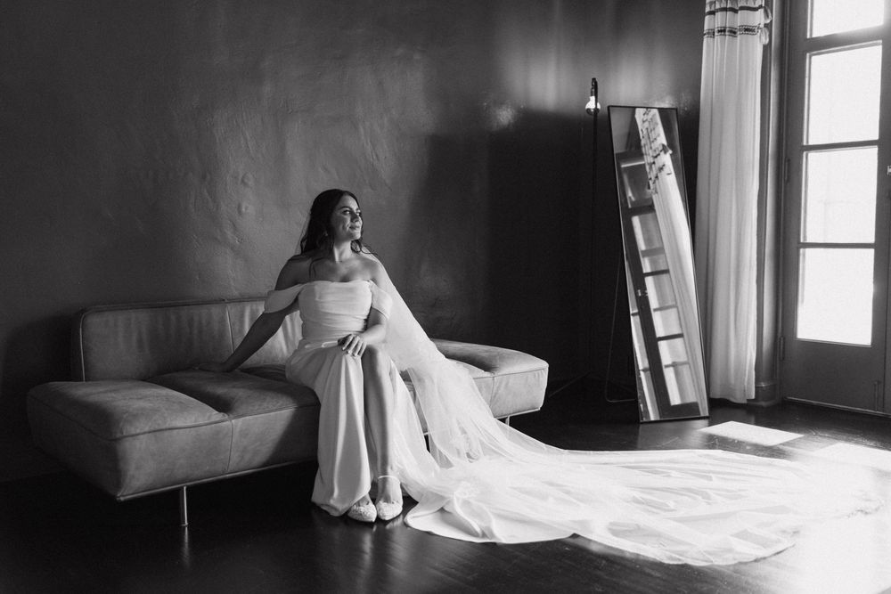 Black and white photo of bride in white wedding gown seated on sofa gazing out bright window.