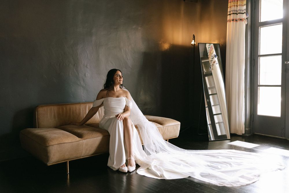 Bride in off-shoulder white wedding dress with long train sitting on leather sofa by sunlit window.