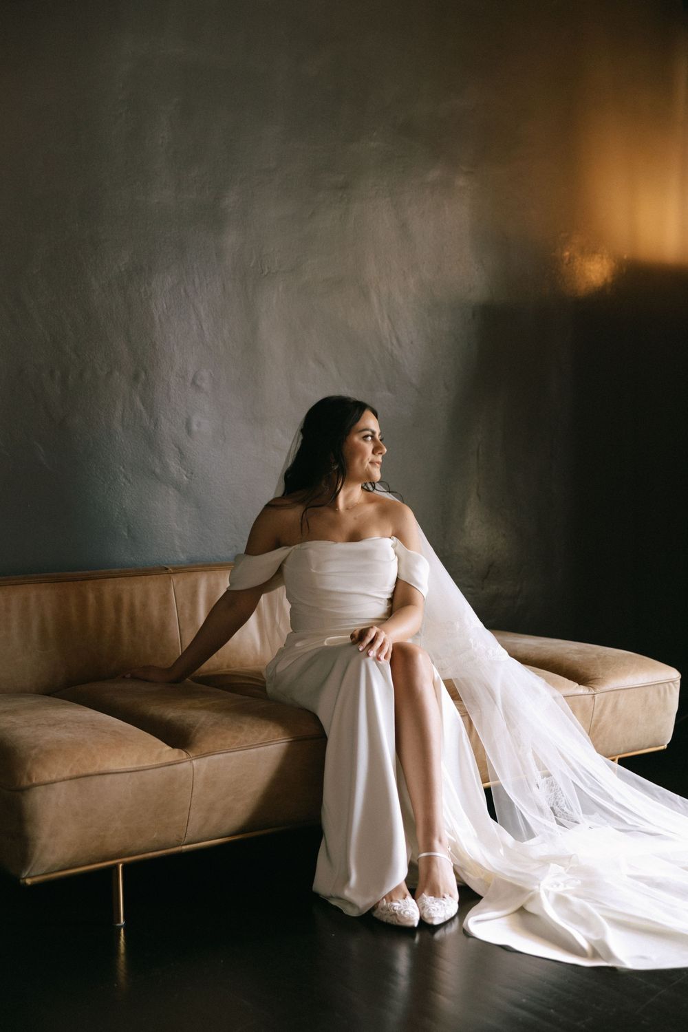 Radiant bride in off-shoulder white gown with veil sitting on tan leather sofa against dark wall.