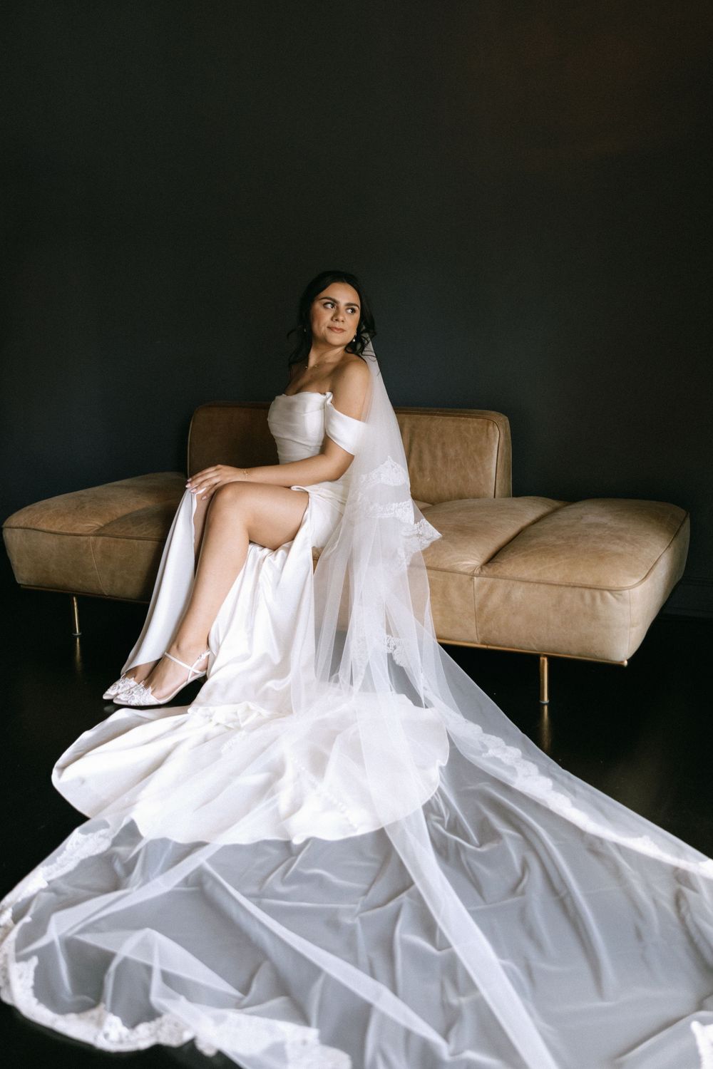 Full-length portrait of bride in strapless white gown with dramatic flowing veil on leather sofa.