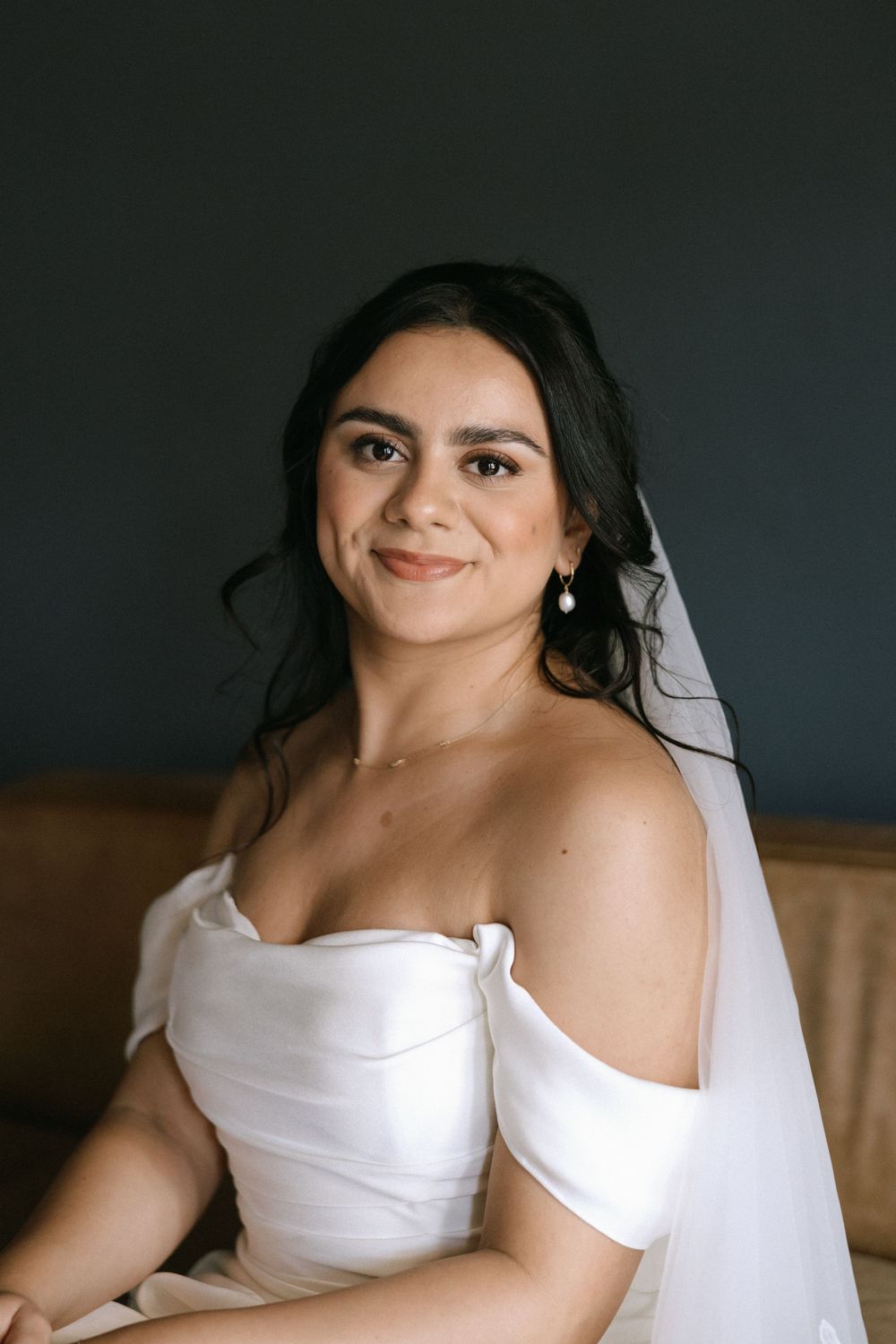 Close-up portrait of smiling dark-haired bride in off-shoulder white dress wearing pearl earrings.