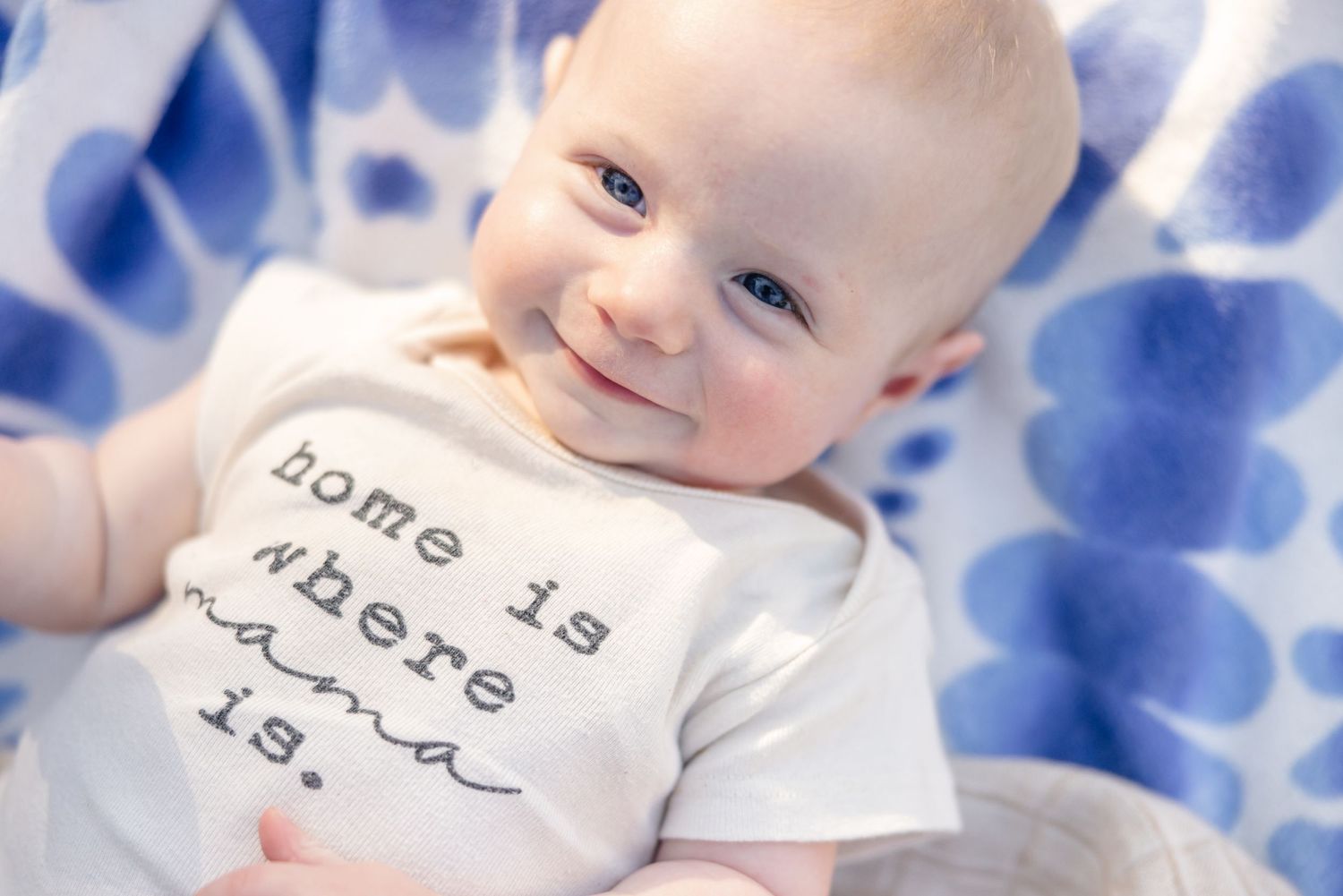 Baby wearing white onesie with text lying on blue and white patterned blanket.