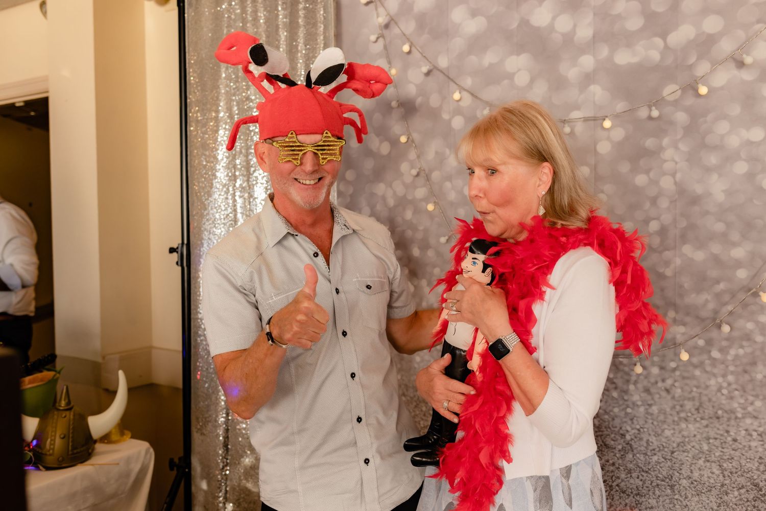 Two people wearing festive red holiday accessories smile while posing together against a sparkly backdrop.