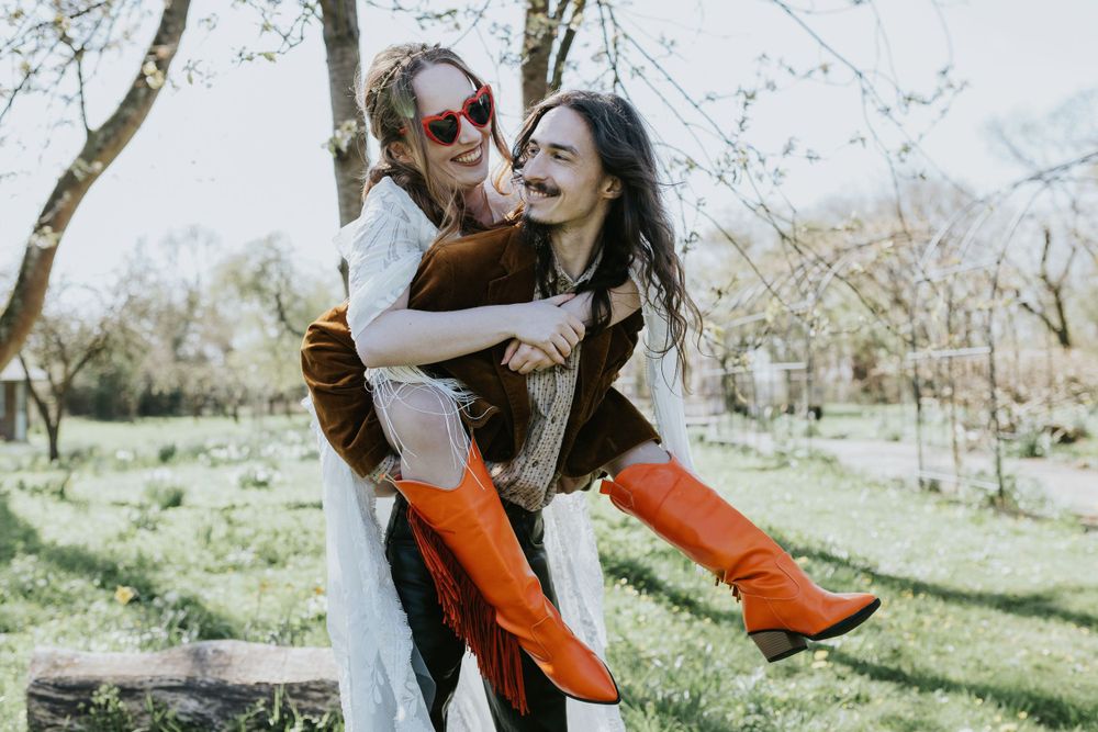 A playful couple embraces outdoors on a sunny day in a garden setting, with bright orange boots adding a pop of color.