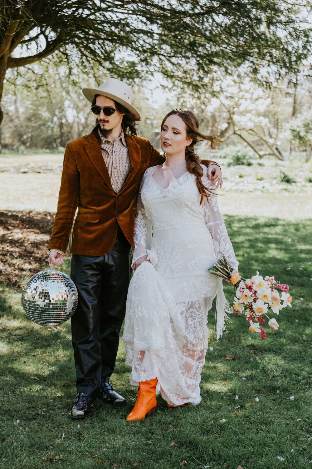 A couple walks outdoors in wedding attire, with a disco ball and bouquet, featuring orange boots and a brown velvet jacket.