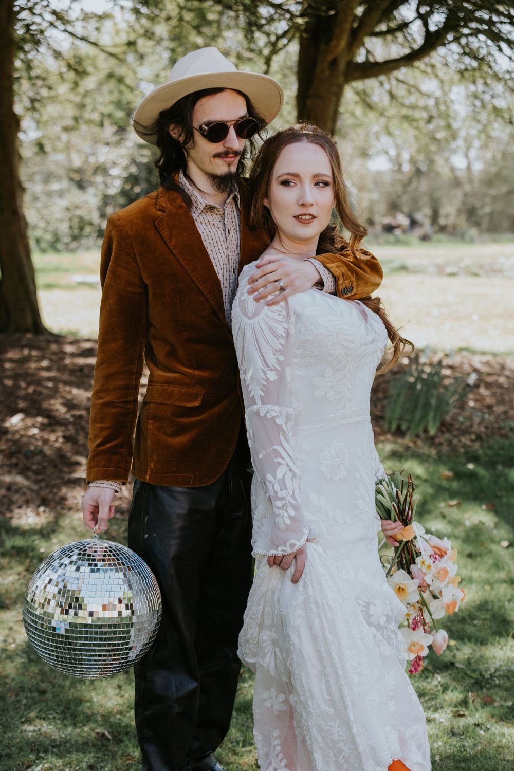 A couple poses in vintage-inspired wedding attire with a disco ball in a garden setting.