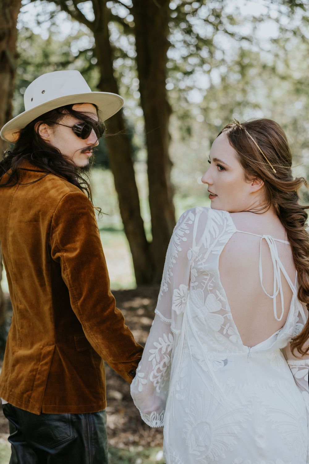 A couple in wedding attire stands in a forest setting, with the groom wearing a brown suede jacket and cowboy hat.