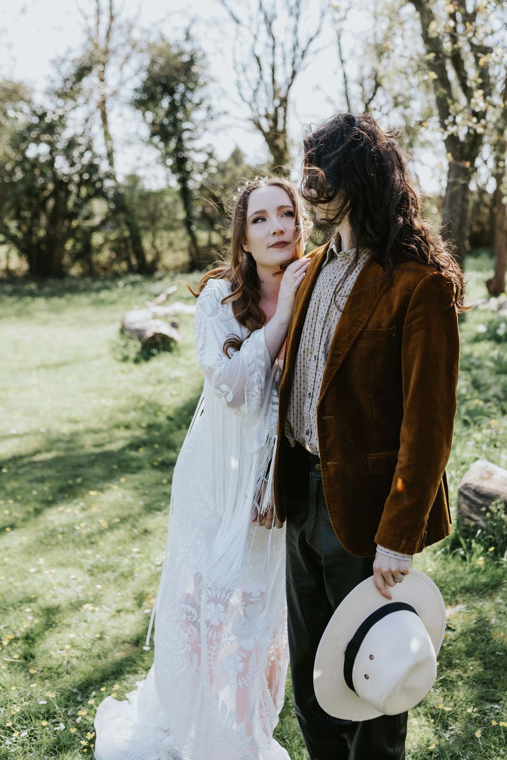 A romantic couple in vintage-inspired attire standing together in a sunlit garden with a white hat.