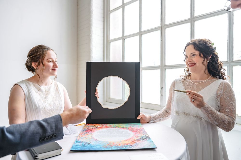 Two brides in white dresses hold a black frame with circle cutout at a wedding signing ceremony indoors.